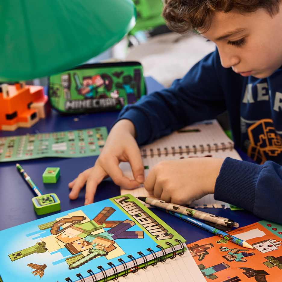 Dečak piše u svesci sa temom video igre, okružen olovkama i pernicom.