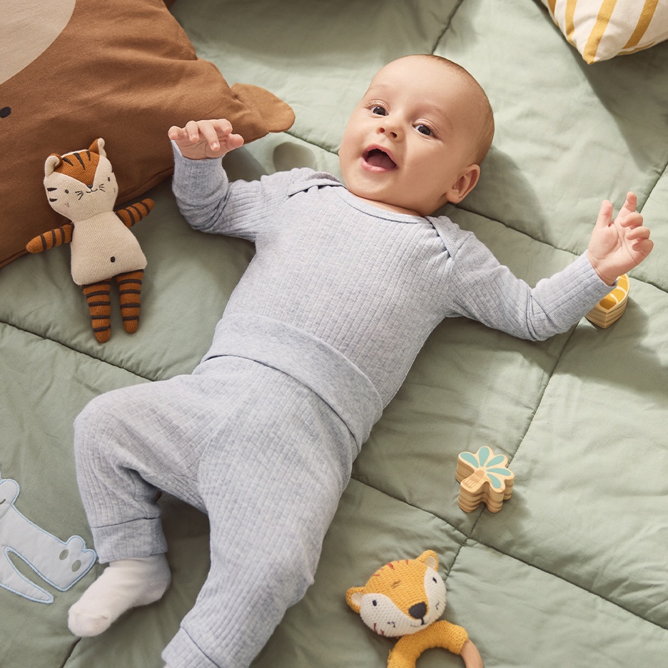 Beba u svetloplavom rebrastom kompletu leži na zelenom jorganu sa plišanim igračkama.