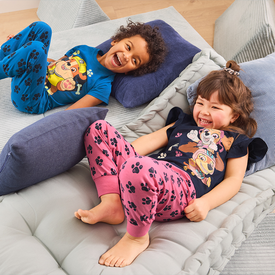 Dvoje dece u pidžamama sa motivima šapa i crtanih junaka, leže i smeju se na podnim jastucima.