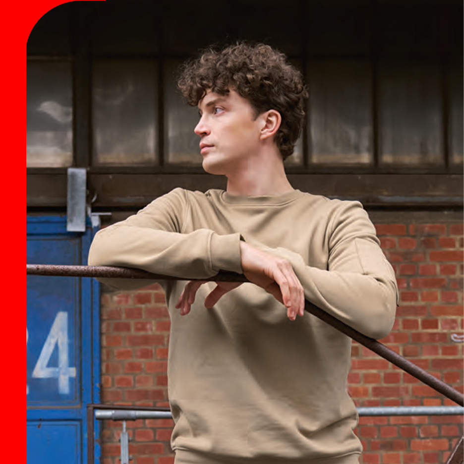 Muškarac u bež duksu naslonjen na metalnu šipku, sa ciglenim zidom u pozadini.