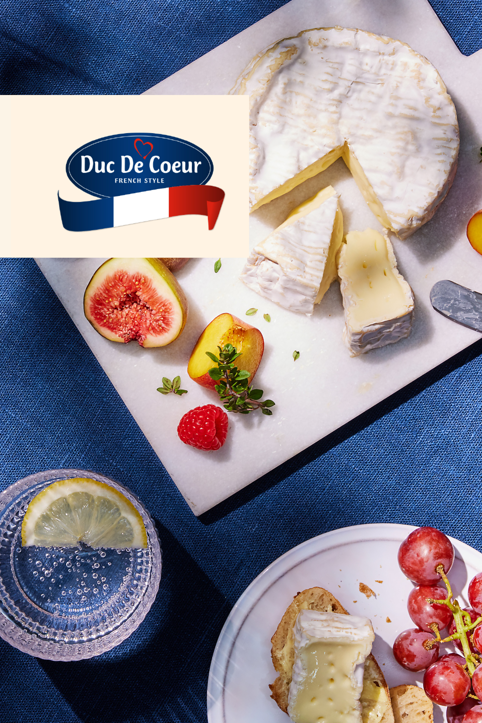 Kremasti sir, voće i piće na plavoj pozadini, sa logotipom „Duc De Coeur French Style“.