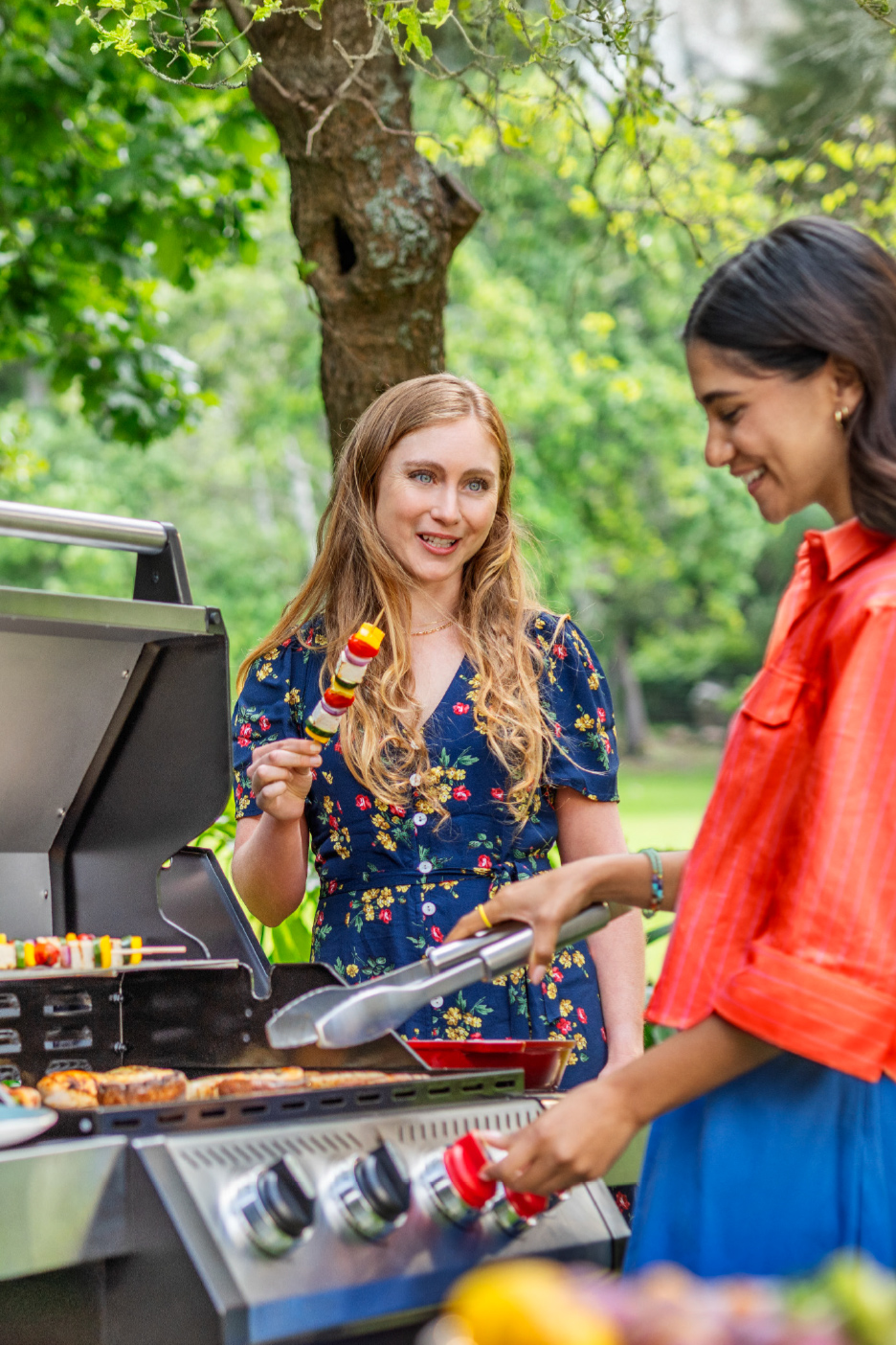 Dve žene roštiljaju u bašti, jedna drži ražnjić, druga okreće hranu kleštima.