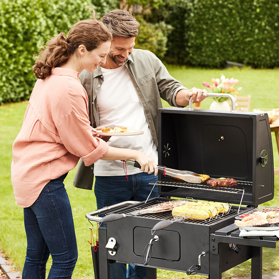 Par roštilja u bašti, žena okreće hranu na roštilju, muškarac drži tanjir.