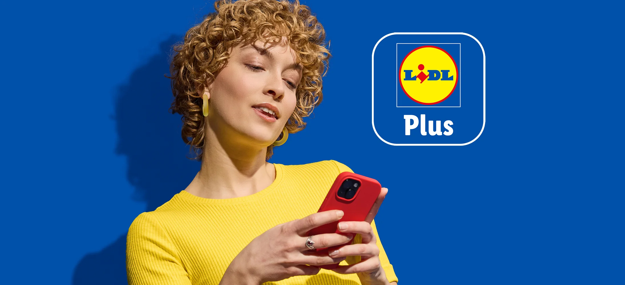 Žena sa kovrdžavom kosom drži crveni telefon, pored Lidl Plus logotipa na plavoj pozadini.