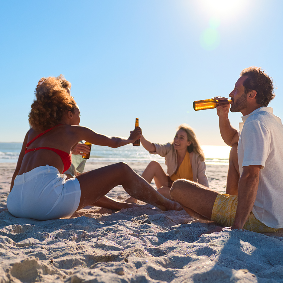 Prijatelji uživaju u pivu na plaži, sunce sija, a more je u pozadini.