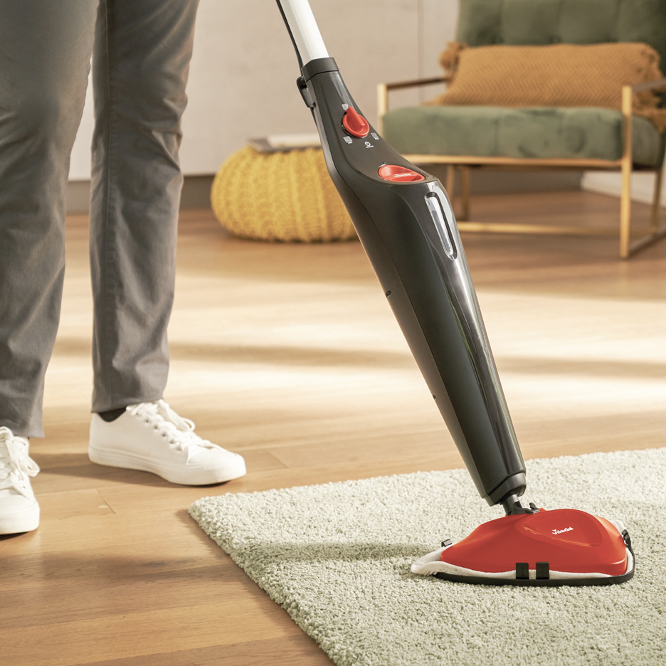 Osoba čisti pod parnim čistačem, sa drvenim podom i zelenim tepihom.