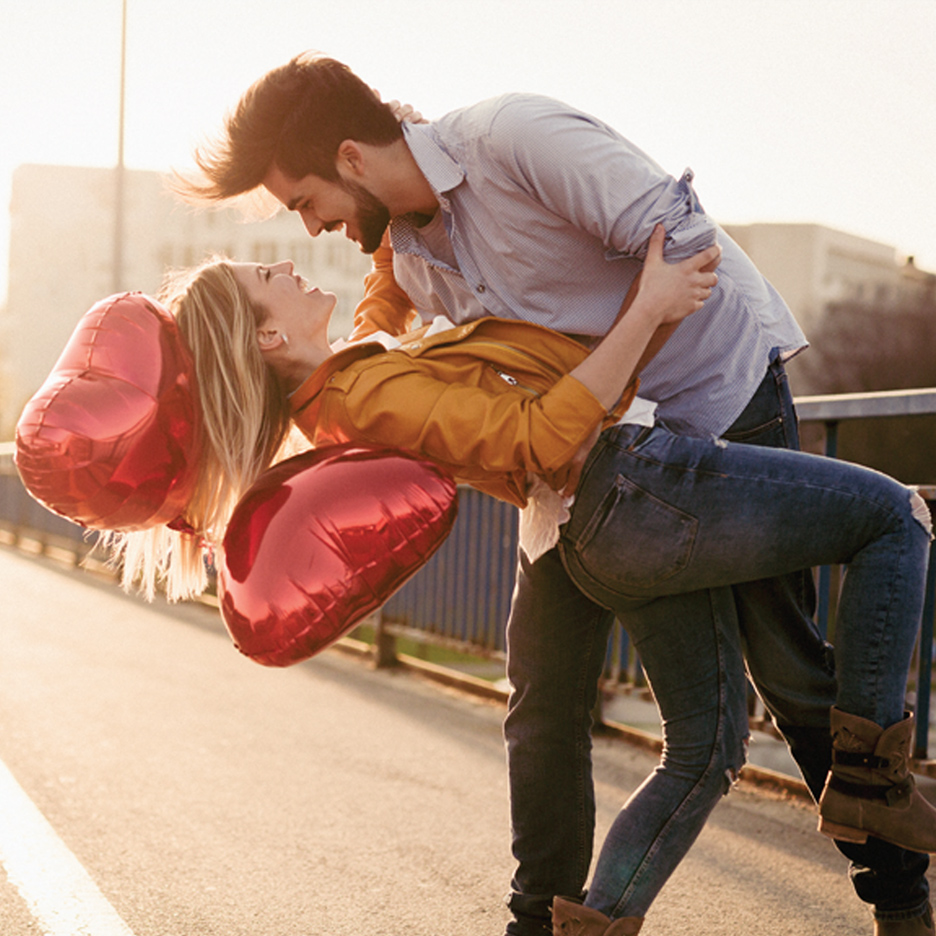 Par se grli na otvorenom, žena drži crvene balone u obliku srca.