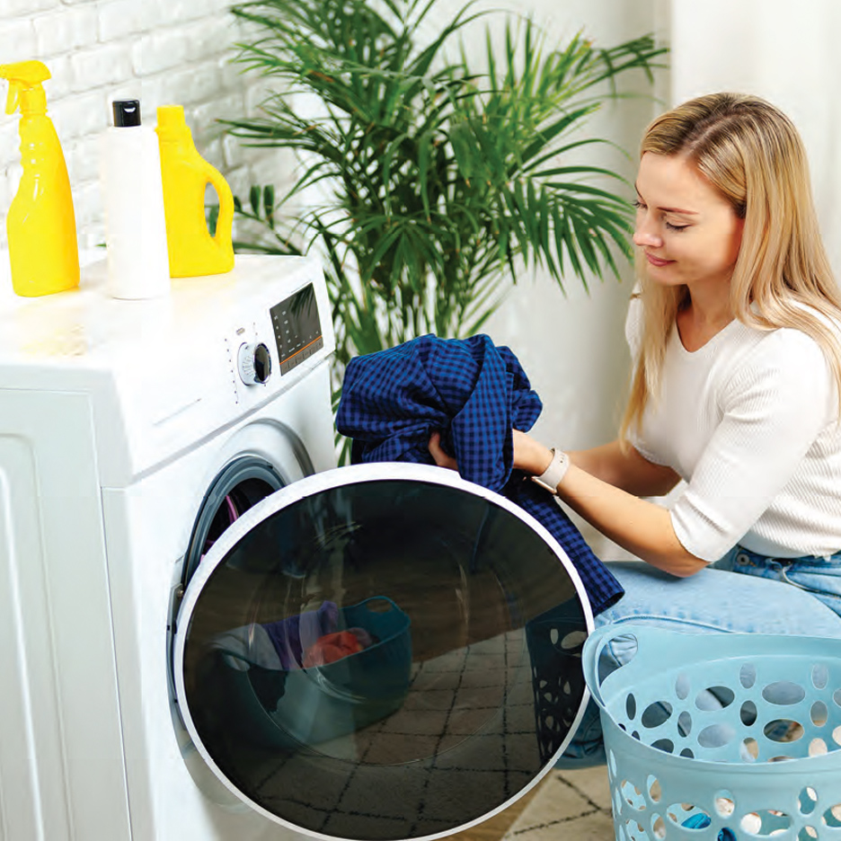 Žena stavlja plavu kariranu košulju u veš mašinu pored korpe za veš.