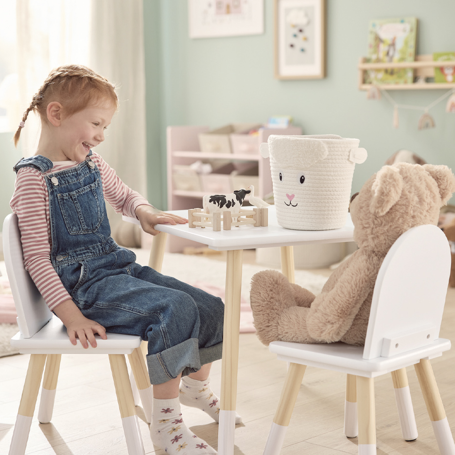 Devojčica i plišani meda sede za belim drvenim stolom sa igračkama i korpom u obliku ovce.