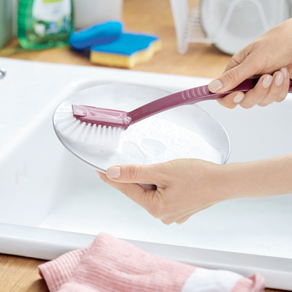 Žena pere tanjir četkom za sudove u sudoperi, sa deterdžentom i sunđerima u pozadini.